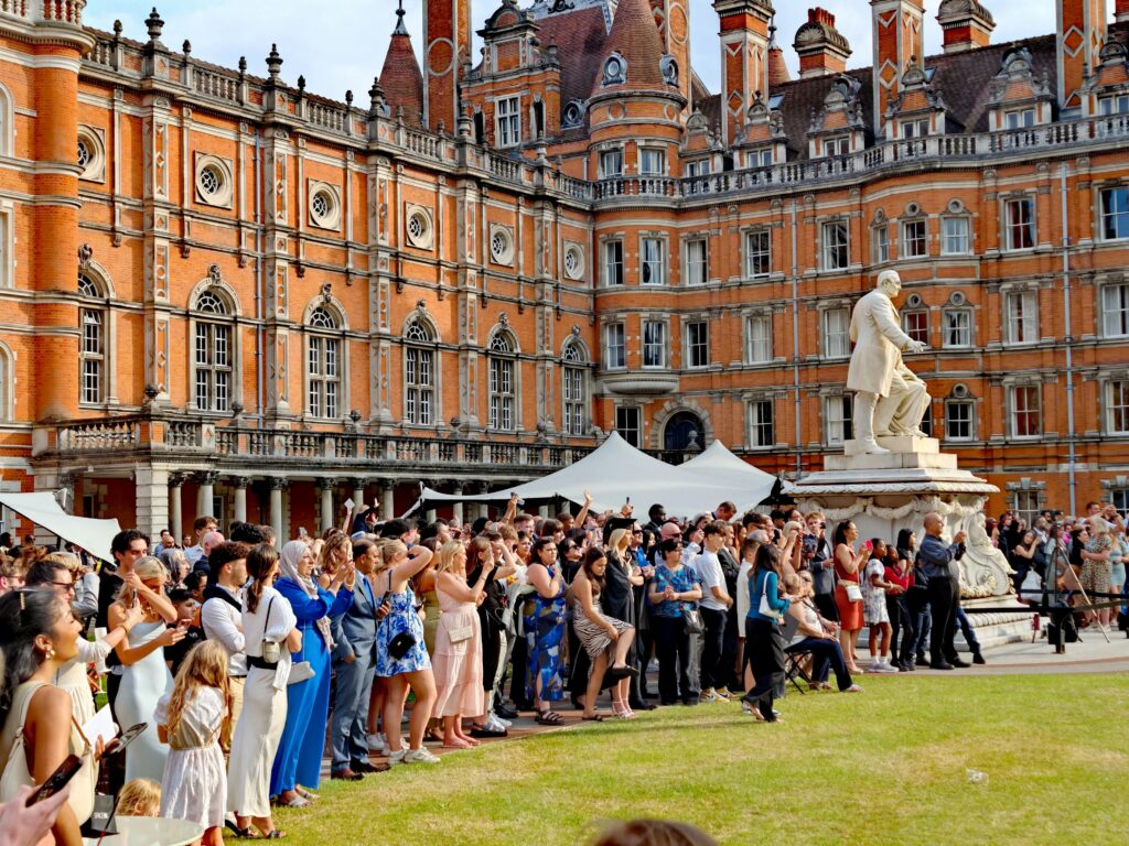 A large crowd gathers for a graduation ceremony at a historic university in England, featuring ornate architecture.